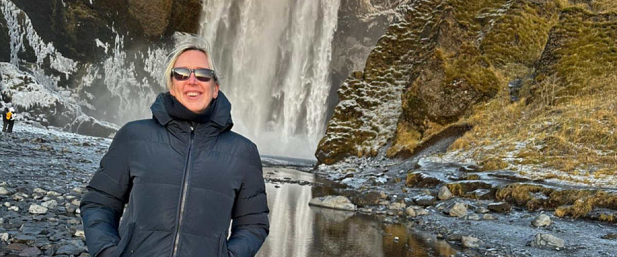 Een schoolleider staat warm aangekleed voor de Skógafoss-waterval in IJsland, op een winterse ochtend met laag zonlicht. Ze draagt een donkere winterjas, jeans en laarzen en glimlacht terwijl ze midden op de rotsige, natte ondergrond poseert. De indrukwekkende waterval en het ruige IJslandse landschap op de achtergrond verwijzen naar haar studiereis, waarin ze inzichten opdeed over inclusief onderwijs, burgerschap en de hechte gemeenschap rond IJslandse scholen.