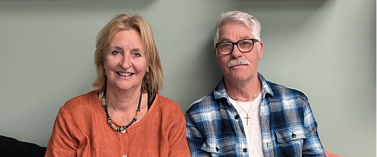 Pensionado's Juf Yvonne en meester Aad zitten ontspannen lachend naast elkaar op een bank, in een huiselijke sfeer. Ze kijken recht in de camera.