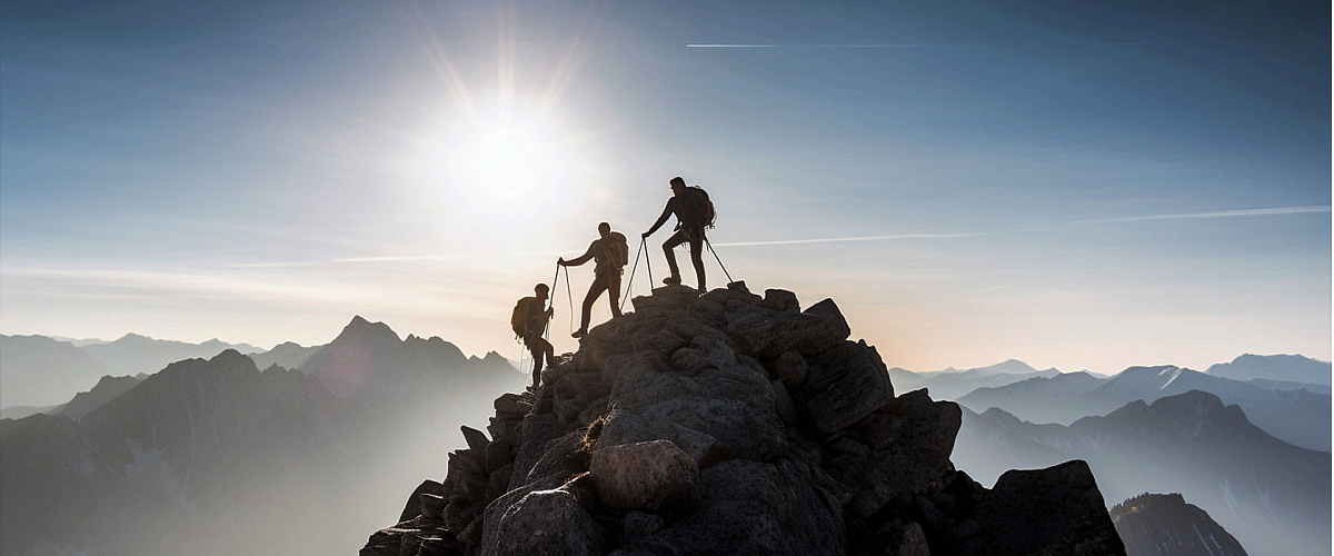Drie klimmers op de top van een bergkam, gesilhouetteerd tegen de felle zon. Dit beeld van een gedeelde klim symboliseert de 'expeditie' van Librijn naar de toekomst, waarbij de route nog niet vastligt en alle stemmen meetellen bij het bepalen van de 'stip op de horizon'.