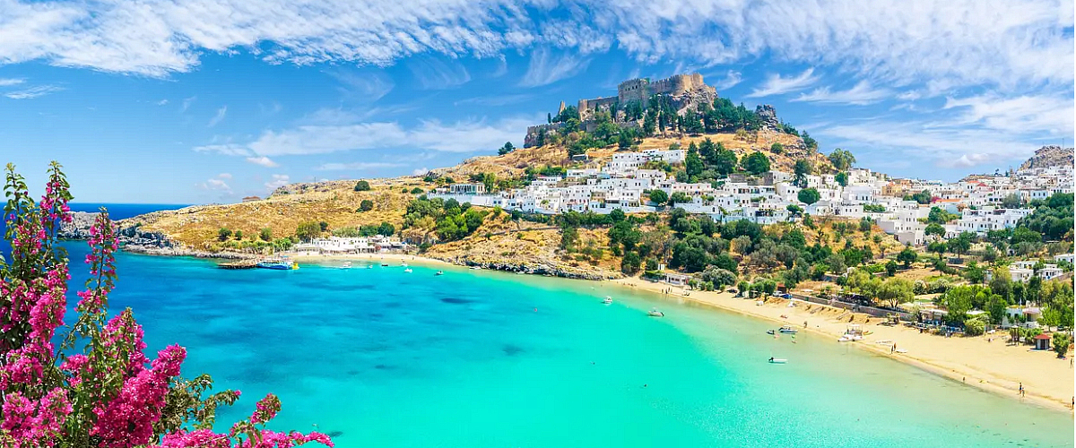 Uitzicht op de turquoise baai van Lindos op Rhodos, met witte huisjes tegen een heuvel, een historisch fort bovenop en felroze bougainville in de voorgrond onder een heldere blauwe lucht.